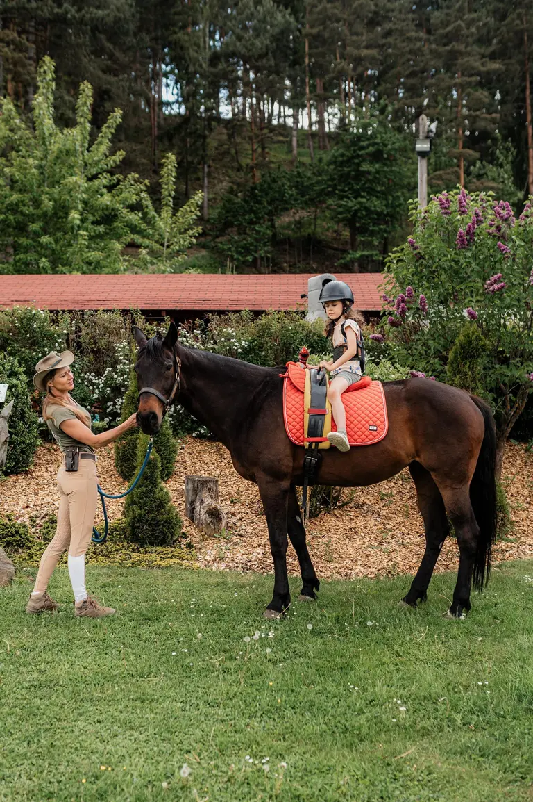Žena vedie koňa, na ktorom sedí dieťa s prilbou vo Villa Betula Resort.
